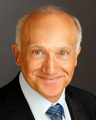 Mann in dunklem Anzug und wei&szlig;em Hemd mit blauer Krawatte vor dunklem Hintergrund l&auml;chelt freundlich in die Kamera