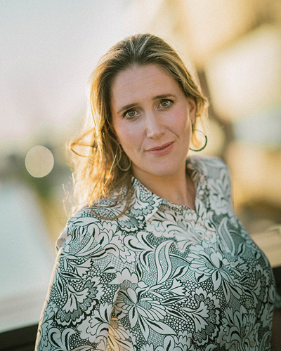 Frau in Hemd mit floralem Muster schaut freundlich in die Kamera w&auml;hrend die Sonne ihre Haare von hinten erstrahlen l&auml;sst